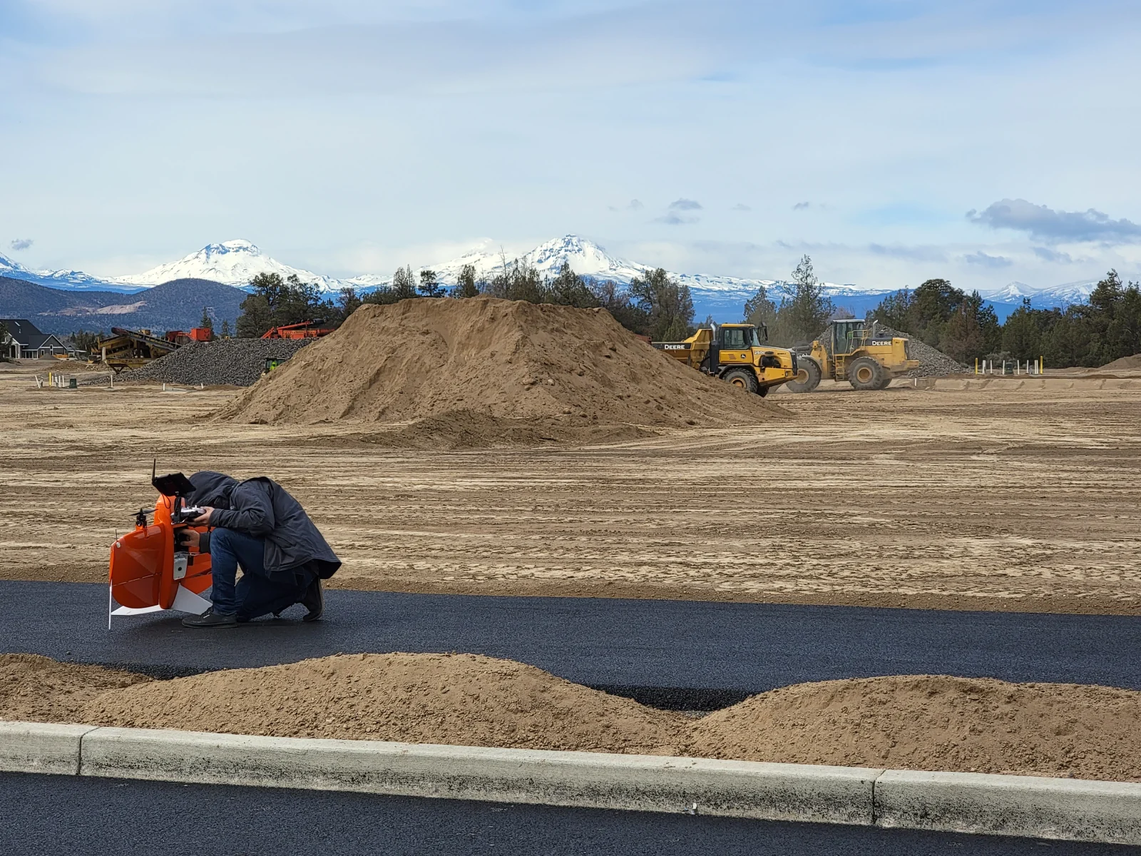 Drone operator preparing survey equipment at a construction site, with earthmoving equipment and mountains behind